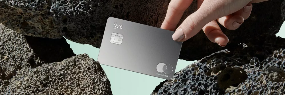 Main tenant une carte bancaire N26 en métal posée sur des roches sombres, design épuré et haut de gamme