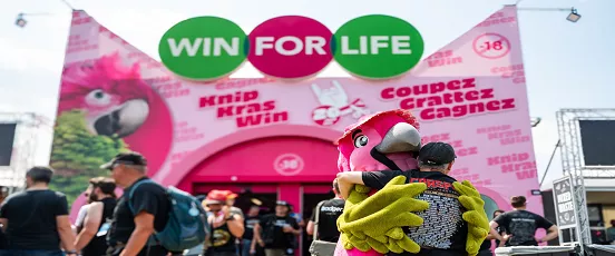 Stand Win For Life au festival belge avec mascotte et festivaliers devant l’animation Coupez, Grattez, Gagnez